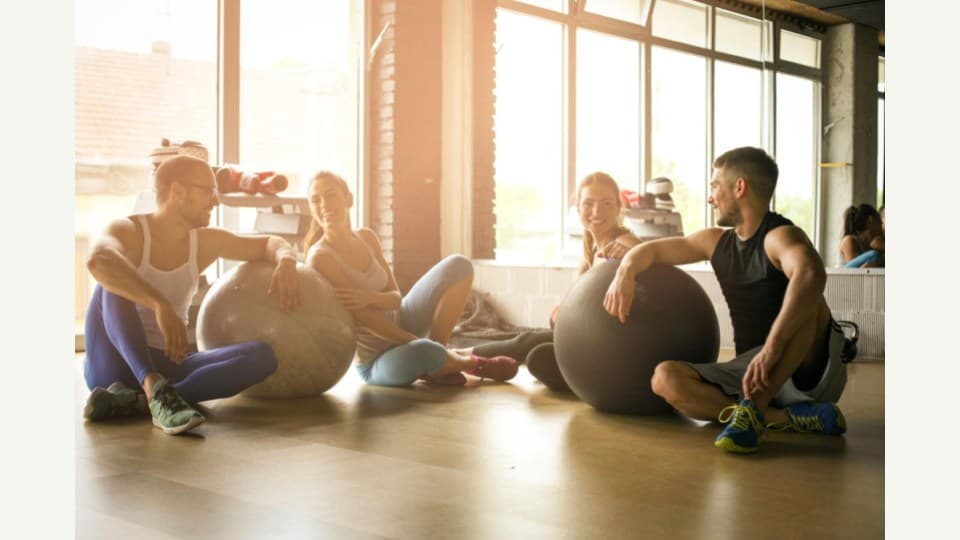 Cuatro amigos descansando con pelotas de pilates en un gimnasio luminoso al atardecer — comunidad y cuidado integral